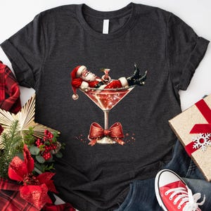 May include: A dark gray t-shirt with a festive design featuring Santa Claus relaxing in a martini glass. The glass is filled with a red liquid and has a sparkly rim. Santa is wearing a red suit with white trim and a white beard. He is holding a martini glass in one hand and has a leg resting on the rim of the glass.