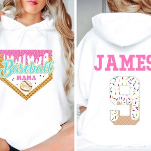 K&ouml;nnte beinhalten: Wei&szlig;er Hoodie mit einem rosa und blauen Baseball-Motiv auf der Vorderseite, auf dem "Baseball Mama" steht, und einer pinkfarbenen Nummer 9 mit Streuseln auf der R&uuml;ckseite, auf der "JAMES" steht.