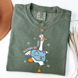 K&ouml;nnte beinhalten: Ein gr&uuml;nes T-Shirt mit einer Comic-Gans, die eine Krankenschwesteruniform und ein Stethoskop tr&auml;gt. Der Text "Registered Silly Goose" ist auf dem T-Shirt aufgedruckt.