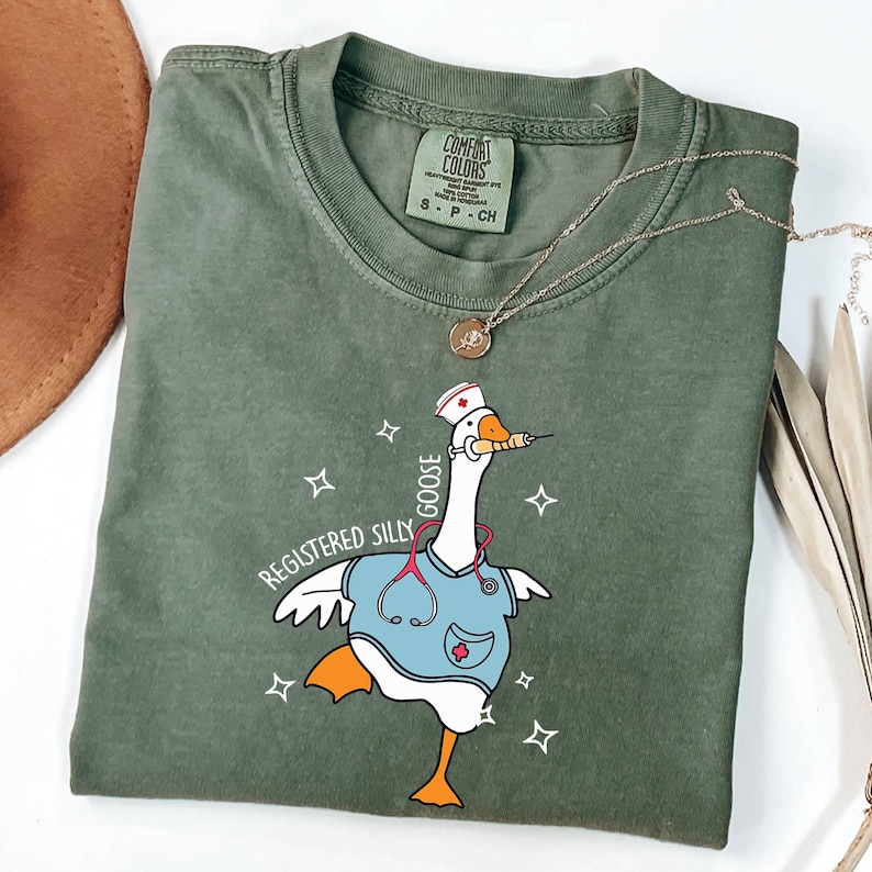 K&ouml;nnte beinhalten: Ein gr&uuml;nes T-Shirt mit einer Comic-Gans, die eine Krankenschwesteruniform und ein Stethoskop tr&auml;gt. Der Text "Registered Silly Goose" ist auf dem T-Shirt aufgedruckt.