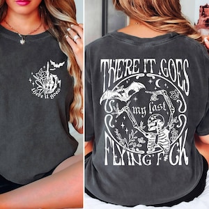 K&ouml;nnte beinhalten: Ein dunkelgraues T-Shirt mit einem wei&szlig;en Grafikdruck eines Skeletts, das eine Fledermaus h&auml;lt, und dem Text "There it goes my last flying f*ck".