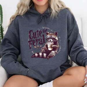 May include: A dark gray hoodie with a raccoon graphic and the text "Cute But Feral" printed on the front. The raccoon is holding a coffee cup.