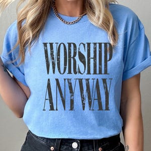 May include: A light blue t-shirt with the words "WORSHIP ANYWAY" printed in large, distressed black letters. The shirt has short sleeves and a crew neck. The model is wearing a gold chain necklace and dark blue jeans.