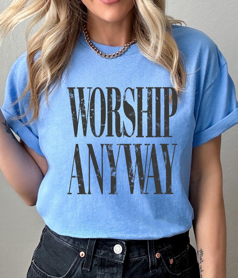 May include: A light blue t-shirt with the words "WORSHIP ANYWAY" printed in large, distressed black letters. The shirt has short sleeves and a crew neck. The model is wearing a gold chain necklace and dark blue jeans.