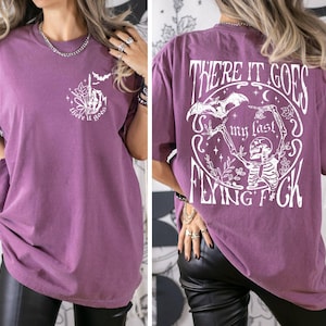 K&ouml;nnte beinhalten: Ein lilafarbenes Oversize-T-Shirt mit einem wei&szlig;en Grafikdruck eines Skeletts, das eine Fledermaus mit dem Text "There it goes my last flying f*ck" h&auml;lt.