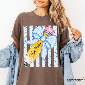 May include: A brown short-sleeve t-shirt featuring a graphic of a yellow pencil with a pink eraser and a large light blue bow with white hearts. The pencil has the text Mrs. Miller written on it, set against a background of light blue vertical stripes.