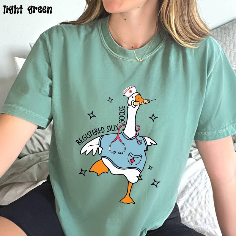 K&ouml;nnte beinhalten: Ein hellgr&uuml;nes T-Shirt mit einer Cartoon-Gans, die eine Krankenschwesterm&uuml;tze und einen Kittel tr&auml;gt. Die Gans h&auml;lt eine Spritze und der Text "Registered Silly Goose" ist auf dem T-Shirt aufgedruckt.