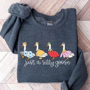 May include: Dark gray sweatshirt featuring four cartoon geese dressed in whimsical outfits, including a floral dress, a yellow cape, a strawberry costume, and a pink flower collar. The text "just a silly goose" is written in cursive below the geese.