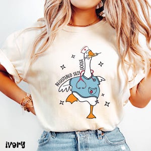 K&ouml;nnte beinhalten: Ein wei&szlig;es T-Shirt mit einer Cartoon-Gans, die eine Krankenschwesteruniform und ein Stethoskop tr&auml;gt. Die Gans h&auml;lt eine Spritze im Schnabel. Der Text "Registered Silly Goose" ist auf dem Shirt aufgedruckt.