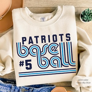May include: Cream-colored sweatshirt with the words "PATRIOTS BASEBALL #5" in navy and sky blue. The design features a retro font and striped accents. The sweatshirt is folded and placed on a wooden surface.
