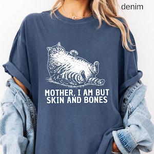 以下が含まれることがあります： デニムブルーのTシャツ。白いイラストはリラックスした猫。テキスト「MOTHER, I AM BUT SKIN AND BONES」が下に印刷されています。「denim」の文字が右上隅に印刷されています。