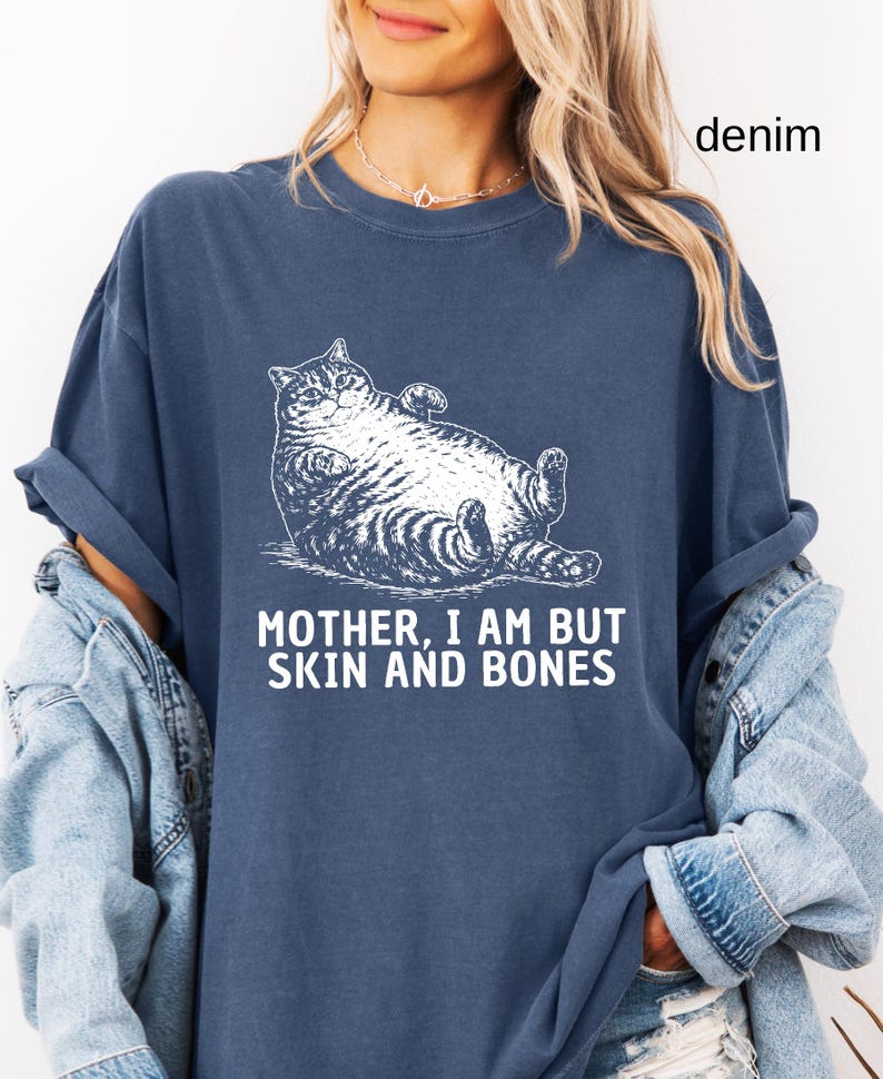 以下が含まれることがあります： デニムブルーのTシャツ。白いイラストはリラックスした猫。テキスト「MOTHER, I AM BUT SKIN AND BONES」が下に印刷されています。「denim」の文字が右上隅に印刷されています。