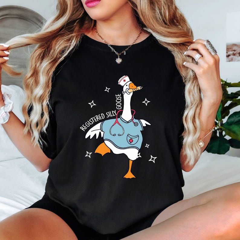 K&ouml;nnte beinhalten: Schwarzes T-Shirt mit einer Cartoon-Gans, die eine Krankenschwesterm&uuml;tze und einen Kittel tr&auml;gt. Der Text "Registered Silly Goose" ist auf dem Shirt aufgedruckt.