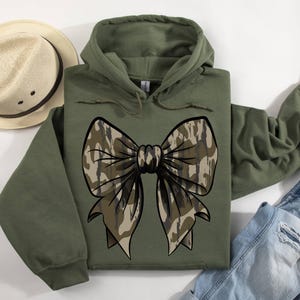 May include: A green hooded sweatshirt with a large camouflage bow on the front. The bow is made of a brown and tan camouflage pattern.