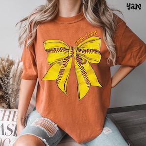 May include: Burnt orange t-shirt featuring a large yellow bow design with red stitching, resembling a softball. The text "YOUR TEAM" is arched across the top of the bow. The shirt has short sleeves and a relaxed fit.