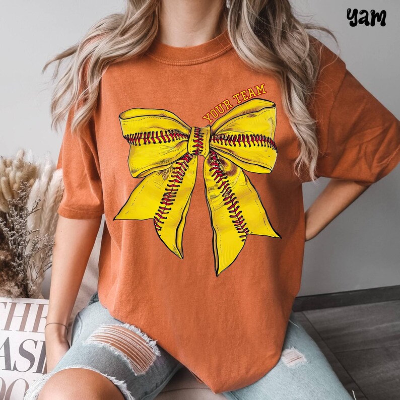 May include: Burnt orange t-shirt featuring a large yellow bow design with red stitching, resembling a softball. The text "YOUR TEAM" is arched across the top of the bow. The shirt has short sleeves and a relaxed fit.