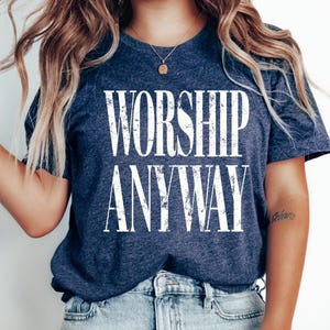 May include: A navy blue t-shirt with the words "WORSHIP ANYWAY" in large, distressed white letters. The shirt is made of a soft material and is paired with light blue denim jeans. A gold necklace is visible.