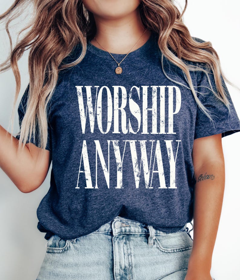 May include: A navy blue t-shirt with the words "WORSHIP ANYWAY" in large, distressed white letters. The shirt is made of a soft material and is paired with light blue denim jeans. A gold necklace is visible.