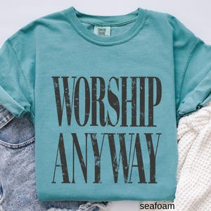 May include: Seafoam green long-sleeve shirt with the words "WORSHIP ANYWAY" printed in distressed black lettering. The shirt is made of a soft material and has a crew neck. The brand "Comfort Colors" is printed on the tag.