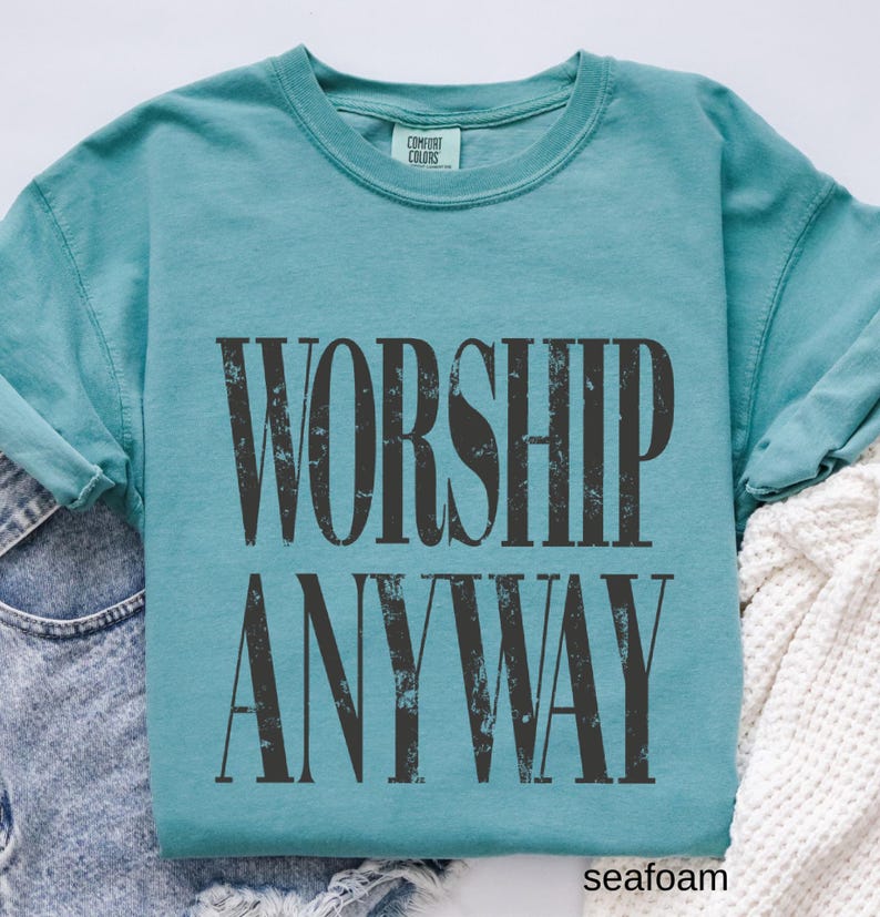 May include: Seafoam green long-sleeve shirt with the words "WORSHIP ANYWAY" printed in distressed black lettering. The shirt is made of a soft material and has a crew neck. The brand "Comfort Colors" is printed on the tag.