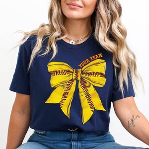 May include: A navy blue t-shirt with a yellow softball bow and the text "YOUR TEAM" printed on the front.