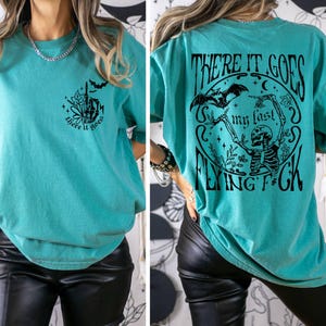 K&ouml;nnte beinhalten: &Uuml;berdimensioniertes T-Shirt in Tealblau mit einem schwarzen Grafikdruck eines Skeletts mit Flederm&auml;usen und dem Text "There it goes my last flying f*ck". Die Vorderseite des Shirts hat einen kleineren Grafikdruck einer Hand mit dem Text "There it goes".