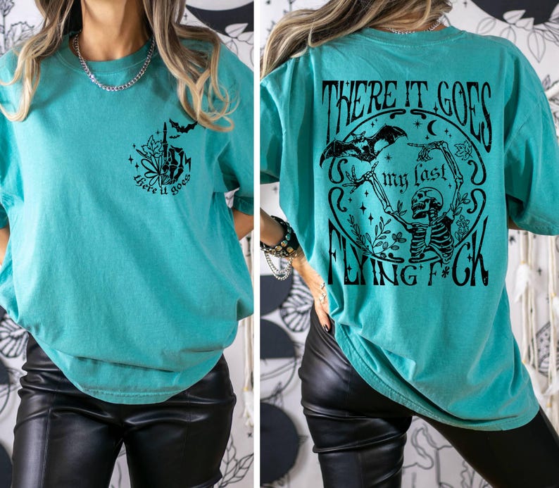 K&ouml;nnte beinhalten: &Uuml;berdimensioniertes T-Shirt in Tealblau mit einem schwarzen Grafikdruck eines Skeletts mit Flederm&auml;usen und dem Text "There it goes my last flying f*ck". Die Vorderseite des Shirts hat einen kleineren Grafikdruck einer Hand mit dem Text "There it goes".