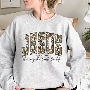 May include: A light gray sweatshirt with the word "JESUS" in large, camouflage-patterned letters. Below the word "JESUS" is the text "JOHN 14:6" and the phrase "the way the truth the life" in a script font. The sweatshirt has long sleeves.