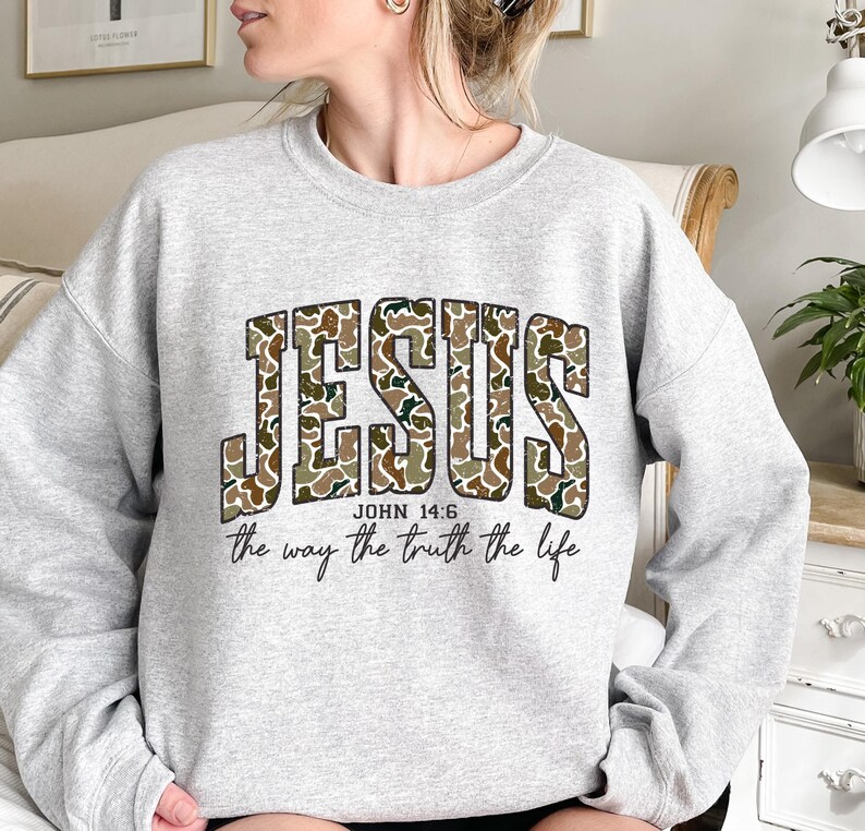 May include: A light gray sweatshirt with the word "JESUS" in large, camouflage-patterned letters. Below the word "JESUS" is the text "JOHN 14:6" and the phrase "the way the truth the life" in a script font. The sweatshirt has long sleeves.