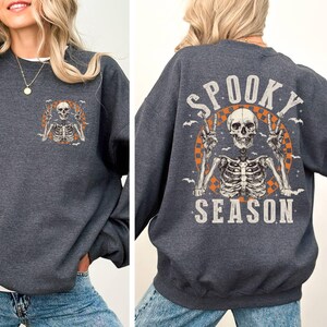 Puede incluir: Una sudadera gris oscura con un gr&aacute;fico de esqueleto y el texto "Spooky Season" impreso en la espalda. El esqueleto est&aacute; levantando dos signos de paz.