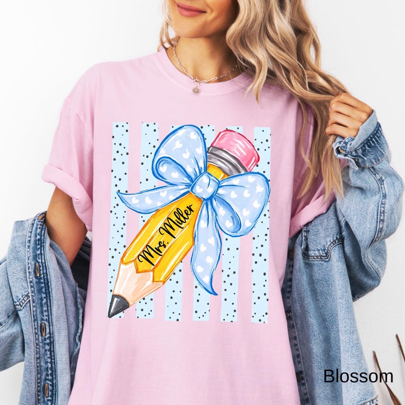 May include: A light pink t-shirt featuring a graphic of a yellow pencil with a pink eraser and a large light blue bow with white hearts. The pencil is inscribed with the text Mrs. Miller and is set against a background of light blue vertical stripes.