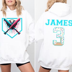 May include: White hoodie featuring a baseball design with crossed bats and a baseball. The back of the hoodie has the name "JAMES" and the number "3" in a teal color with a sprinkle and waffle cone design.