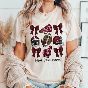 Puede incluir: Una camiseta blanca con un dise&ntilde;o marr&oacute;n que incluye un bal&oacute;n de f&uacute;tbol, un meg&aacute;fono de animadora, un casco de f&uacute;tbol y un lazo. El texto "Cheer" y "Your team name" tambi&eacute;n est&aacute;n incluidos en el dise&ntilde;o.