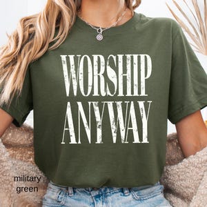 May include: Military green t-shirt with the words "WORSHIP ANYWAY" in large, distressed white letters. The shirt is a crew neck style and appears to be made of a soft material. The text is centered on the front of the shirt.