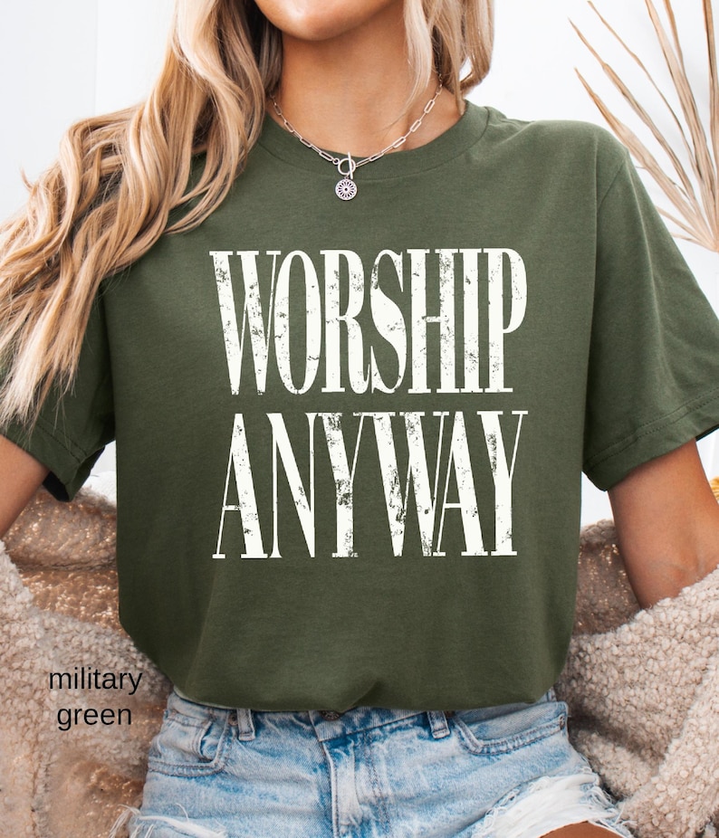 May include: Military green t-shirt with the words "WORSHIP ANYWAY" in large, distressed white letters. The shirt is a crew neck style and appears to be made of a soft material. The text is centered on the front of the shirt.