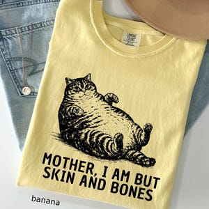 以下が含まれることがあります： バナナ色のTシャツ。黒と白で描かれた、仰向けに寝ている猫のイラストが特徴です。「MOTHER, I AM BUT SKIN AND BONES」の文字が猫の下にプリントされています。「banana」の文字が下にプリントされています。