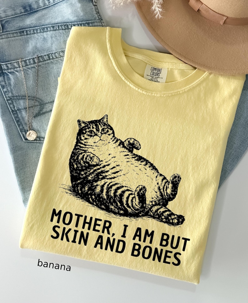 以下が含まれることがあります： バナナ色のTシャツ。黒と白で描かれた、仰向けに寝ている猫のイラストが特徴です。「MOTHER, I AM BUT SKIN AND BONES」の文字が猫の下にプリントされています。「banana」の文字が下にプリントされています。