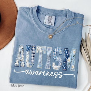 May include: Blue jean Comfort Colors t-shirt with the word "AUTISM" in patchwork letters and the word "awareness" in script. The letters feature floral, gingham, and polka dot patterns. A gold necklace with a pendant is draped on the shirt.