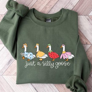 May include: Olive green sweatshirt featuring a whimsical illustration of four geese dressed in various outfits. The geese are wearing a hat, cape, strawberry, and flower skirt. The text "just a silly goose" is written below the geese.