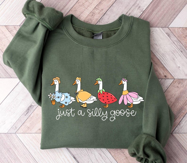 May include: Olive green sweatshirt featuring a whimsical illustration of four geese dressed in various outfits. The geese are wearing a hat, cape, strawberry, and flower skirt. The text "just a silly goose" is written below the geese.
