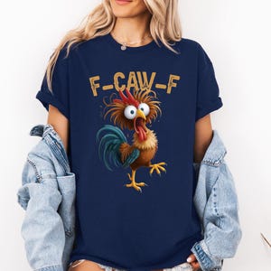 May include: Navy blue t-shirt featuring a cartoon rooster with wide eyes and an open beak. The text "F-CAW-F" is printed above the rooster. The rooster has brown, red, blue, and yellow colors.