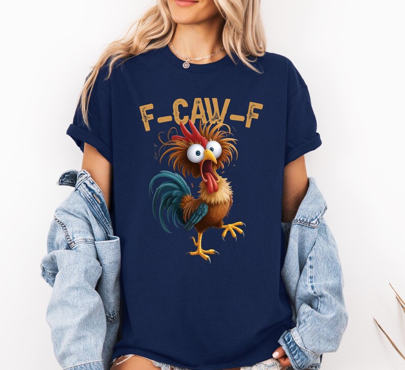 May include: Navy blue t-shirt featuring a cartoon rooster with wide eyes and an open beak. The text "F-CAW-F" is printed above the rooster. The rooster has brown, red, blue, and yellow colors.