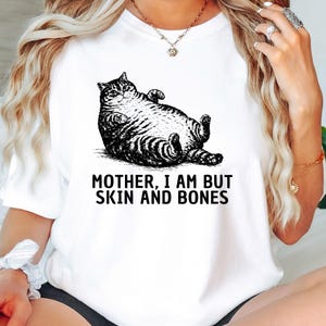 以下が含まれることがあります： 白地のTシャツに、仰向けに寝ている猫の白黒イラストが描かれています。「MOTHER, I AM BUT SKIN AND BONES」の文字が下にプリントされています。普段使いにぴったりのカジュアルなTシャツです。