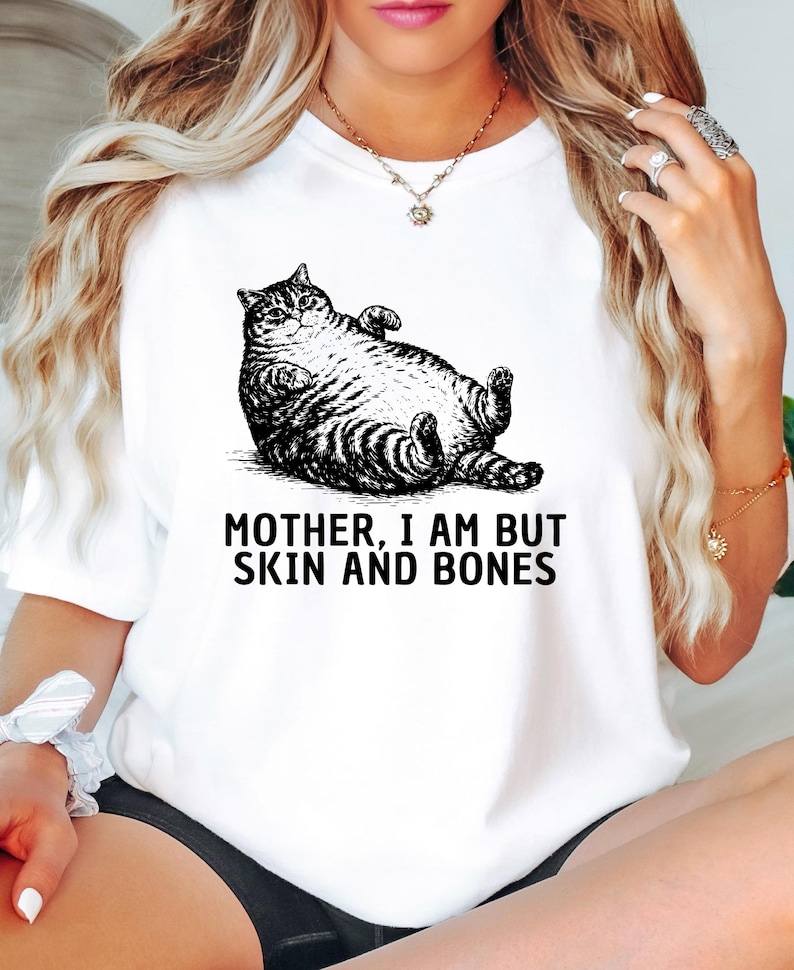以下が含まれることがあります： 白地のTシャツに、仰向けに寝ている猫の白黒イラストが描かれています。「MOTHER, I AM BUT SKIN AND BONES」の文字が下にプリントされています。普段使いにぴったりのカジュアルなTシャツです。