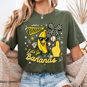 May include: Military green t-shirt with a playful graphic featuring a smiling banana wearing sunglasses, a baseball, and a foam finger. The text "Bananas" and "Let's Go Bananas" are also displayed in yellow.