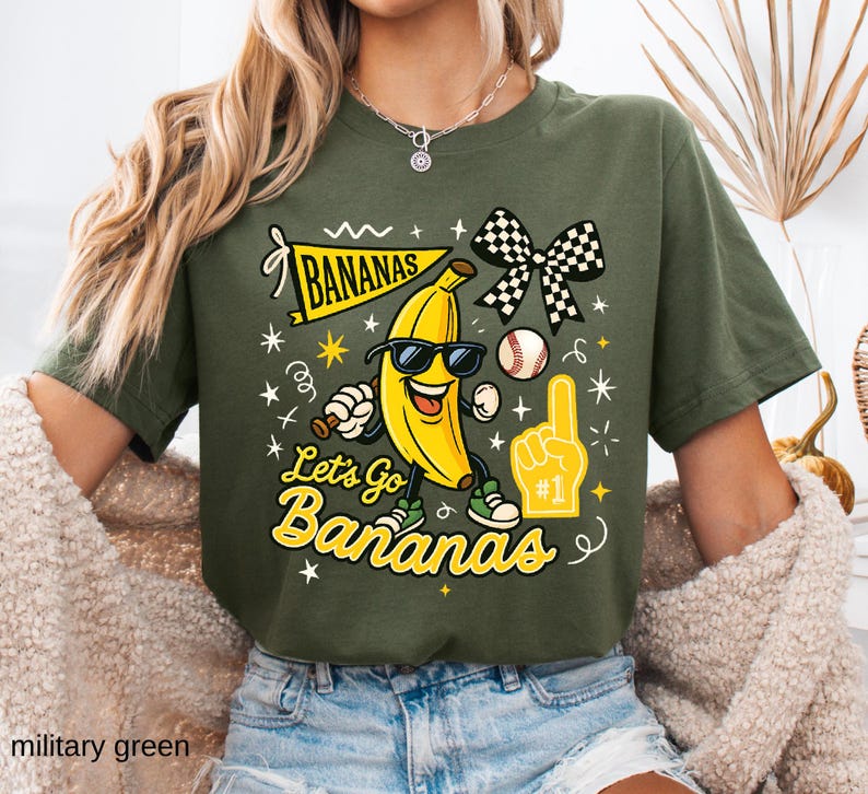 May include: Military green t-shirt with a playful graphic featuring a smiling banana wearing sunglasses, a baseball, and a foam finger. The text "Bananas" and "Let's Go Bananas" are also displayed in yellow.