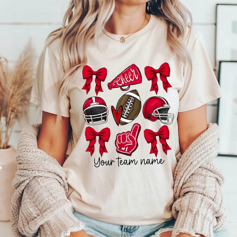 Puede incluir: Una camiseta blanca con un dise&ntilde;o rojo, blanco y negro que presenta un bal&oacute;n de f&uacute;tbol, un meg&aacute;fono de animadora, un coraz&oacute;n, un casco de f&uacute;tbol y una mano con un dedo n&uacute;mero uno. El texto "cheer" y "Your team name" tambi&eacute;n est&aacute;n incluidos en el dise&ntilde;o.