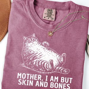 以下が含まれることがあります： ベリー色のTシャツ。リラックスした猫のイラストと「MOTHER, I AM BUT SKIN AND BONES」の文字が特徴です。 金色のネックレスとペンダントがシャツの上にあります。 シャツにはComfort Colorsのタグが付いています。