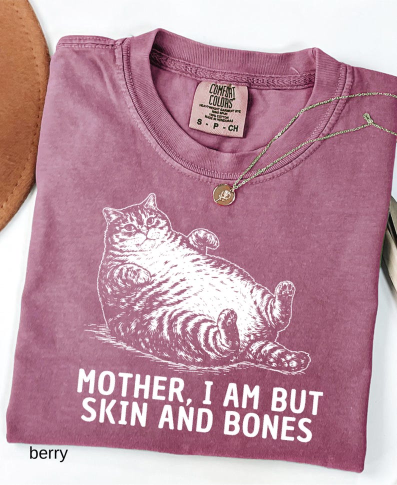 以下が含まれることがあります： ベリー色のTシャツ。リラックスした猫のイラストと「MOTHER, I AM BUT SKIN AND BONES」の文字が特徴です。 金色のネックレスとペンダントがシャツの上にあります。 シャツにはComfort Colorsのタグが付いています。