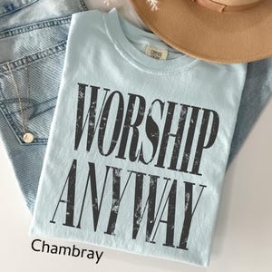 May include: A light blue t-shirt with the words "WORSHIP ANYWAY" in distressed black lettering. The shirt is folded and rests on a pair of blue jeans. A tan hat is visible in the background.